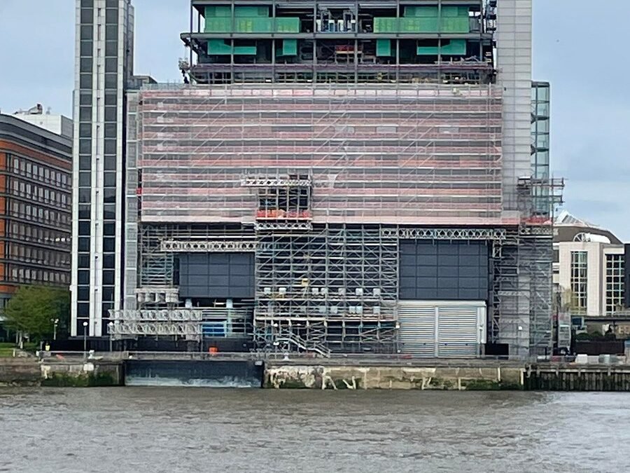 The Telehouse data centre building at Canary Wharf, London, photographed from across a river, showing a large industrial structure covered in scaffolding with satellite dishes mounted on the rooftop