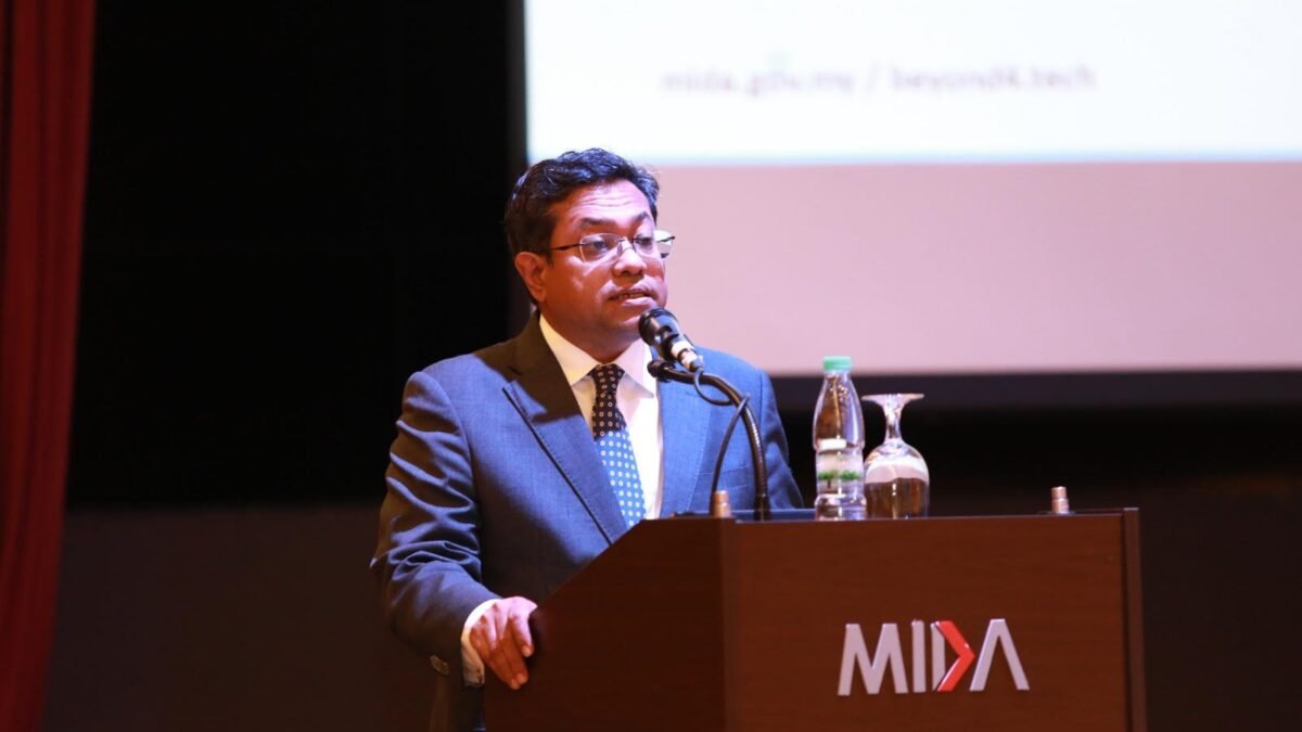 A man in a dark suit and tie speaks at a MIDA-branded wooden podium during the Sarawak Global Supply Chain and Sourcing Programme 2026, with a projected screen visible in the background